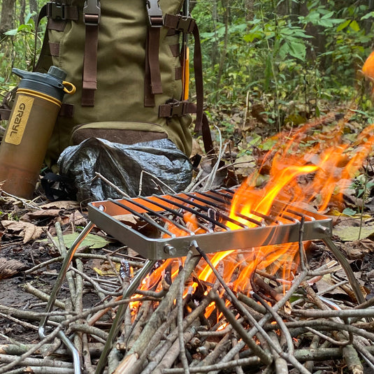 A foldable grill over a small flame with a backpack and Grayl water filtration bottle in the rear
